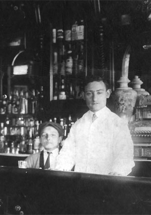 Louis Dumes tending bar.   I'm dating this as 1912 (or before) based on the fact that there is a keg of  Trommer's Beer in the upper right hand corner.  Trommer's Beer was manufactured in Brooklyn and did not have national distribution at that time, so this picture was probably taken in Brooklyn.  We know that by August 1912, Louis was living in Champaign, IL, so this picture must have been taken before that.  

The name of the place, if the cash register isn't lying, is "Reiser's Cafe".  

I've been playing with photoshop and have made a "restored" version of this photo.  The original is <A target=newwin HREF=http://dumes.net/images/louis_dumes_tending_bar.jpg>Here</A>

 (~1912)